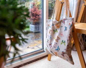 Può includere: Una borsa tote bianca con un motivo a farfalla. La borsa è appesa a una sedia di legno.