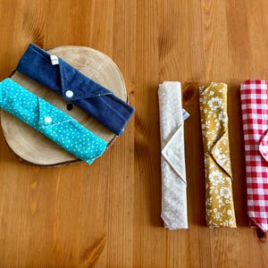 Op de afbeelding: Vijf herbruikbare stoffen servetten, gevouwen en klaar voor gebruik. Twee zijn blauw met witte stippen, één is blauw met witte stiksels, één is beige met een witte bloemenprint en één is rood-wit geruit.