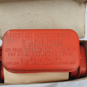 May include: A red metal first aid kit with the text "FIRST AID PACKET, U.S. GOV'T CARLISLE MODEL TO OPEN: PULL TAPE RED COLOR INDICATES BACK OF DRESSING PUT OTHER SIDE NEXT TO WOUND BAUER & BLACK DIV. OF THE KENDALL CO-CHICAGO" printed on the top.