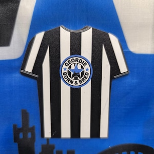May include: Black and white striped shirt with a blue and white circle logo that says "Geordie Born & Bred" with a star and a city skyline inside the circle.