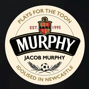 May include: A white circle with a black and gold border. The text "Plays for the Toon" is written in a black circle around the outside of the circle. Inside the circle is a crest with a red and gold shield and the text "EST. 1995". Below the crest is the text "MURPHY" in black with a gold outline. Below the text "MURPHY" is the text "JACOB MURPHY" in black. Below the text "JACOB MURPHY" is a black and white soccer ball with two wheat stalks on either side. The text "Idolised in Newcastle" is written in a black circle around the outside of the circle.