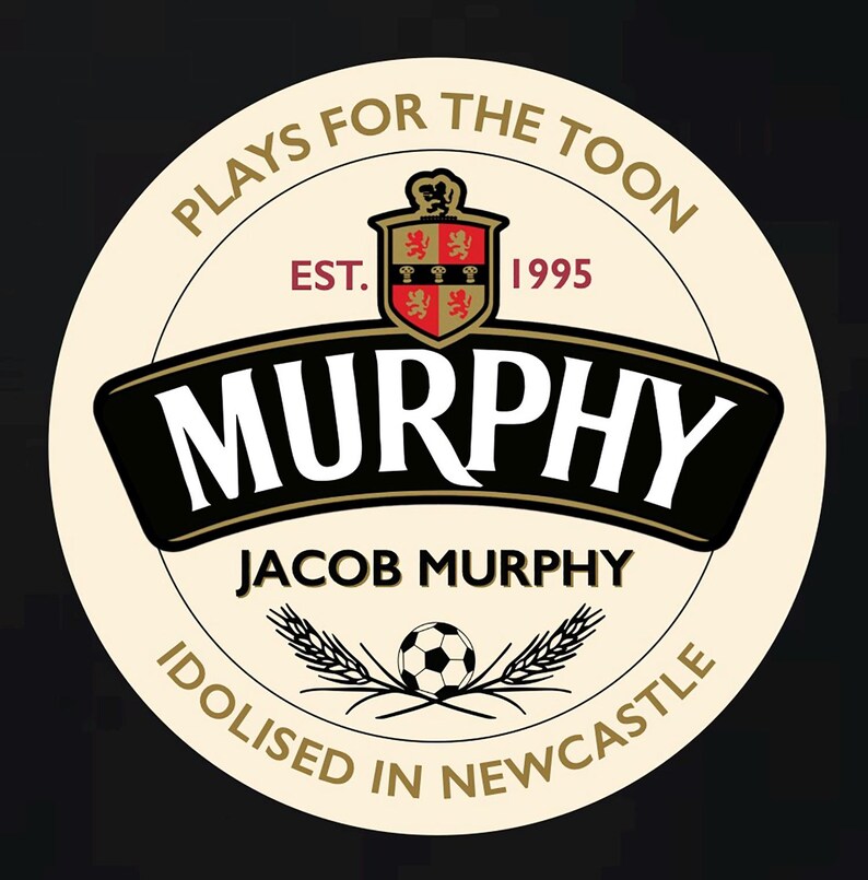 May include: A white circle with a black and gold border. The text "Plays for the Toon" is written in a black circle around the outside of the circle. Inside the circle is a crest with a red and gold shield and the text "EST. 1995". Below the crest is the text "MURPHY" in black with a gold outline. Below the text "MURPHY" is the text "JACOB MURPHY" in black. Below the text "JACOB MURPHY" is a black and white soccer ball with two wheat stalks on either side. The text "Idolised in Newcastle" is written in a black circle around the outside of the circle.