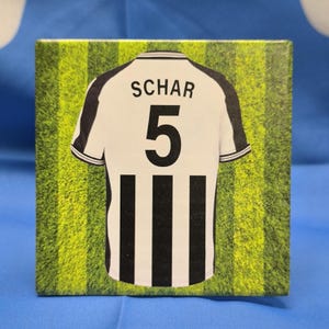 May include: A black and white soccer jersey with the name "SCHAR" and the number "5" on the back. The jersey is on a green and white striped background.