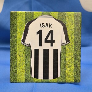 Puede incluir: Una camiseta de f&uacute;tbol a rayas blancas y negras con el nombre "ISAK" y el n&uacute;mero "14" en la espalda. La camiseta est&aacute; sobre un fondo verde y amarillo.