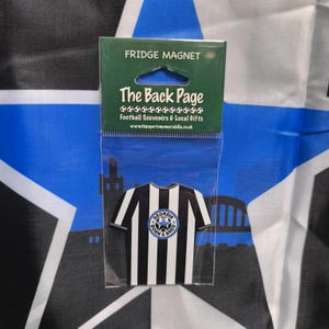 May include: A black and white striped fridge magnet shaped like a soccer jersey with a blue circle logo that says "Geordie Born & Bred". The magnet is packaged in a clear plastic bag with a header that says "Fridge Magnet" and "The Back Page Football Souvenirs & Local Gifts".