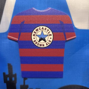 May include: A blue and red striped t-shirt with a white circle logo that says "Newcastle Brown Ale" with a star and a city skyline inside the circle.
