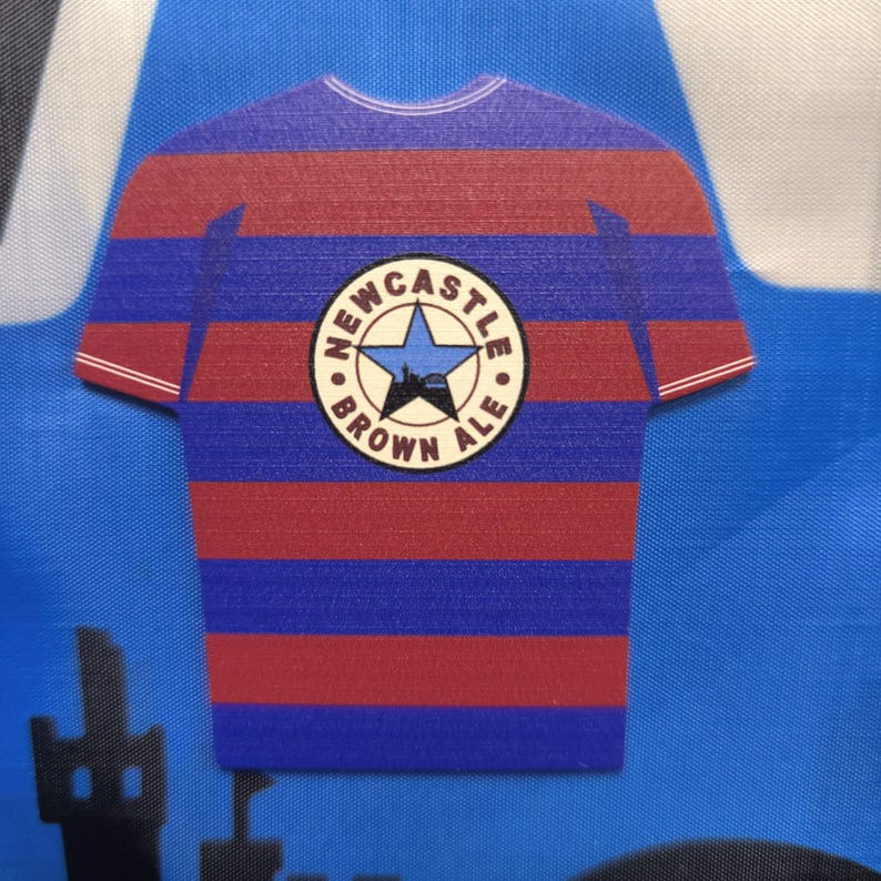 May include: A blue and red striped t-shirt with a white circle logo that says "Newcastle Brown Ale" with a star and a city skyline inside the circle.