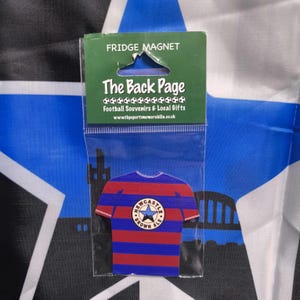 May include: A blue and red striped fridge magnet shaped like a soccer jersey with a white star and the words "Newcastle Brown Ale" on the front. The magnet is packaged in a clear plastic bag with a white label that reads "The Back Page Football Souvenirs & Local Gifts www.thesportsmemorabilia.co.uk"