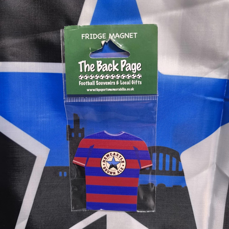 May include: A blue and red striped fridge magnet shaped like a soccer jersey with a white star and the words "Newcastle Brown Ale" on the front. The magnet is packaged in a clear plastic bag with a white label that reads "The Back Page Football Souvenirs & Local Gifts www.thesportsmemorabilia.co.uk"