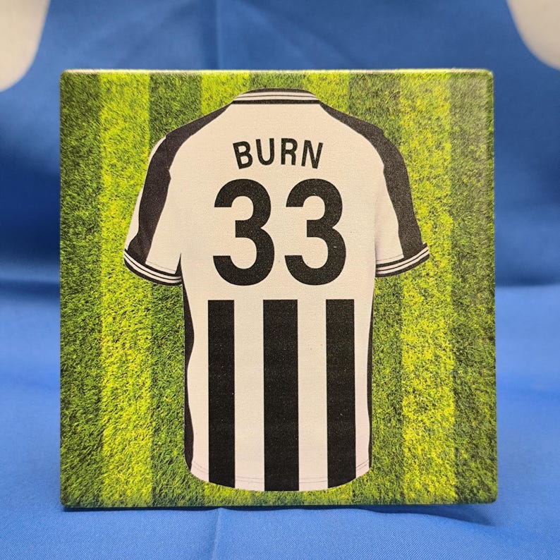 Peut inclure: Un objet d&eacute;coratif carr&eacute; repr&eacute;sentant un maillot de football. Le maillot est blanc avec des rayures et des accents noirs, affichant le nom "BURN" et le num&eacute;ro "33". Le fond est un champ vert avec des rayures verticales.