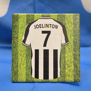K&ouml;nnte beinhalten: Ein schwarz-wei&szlig;es Fu&szlig;balltrikot mit schwarzen und wei&szlig;en Streifen. Das Trikot tr&auml;gt den Namen "JOELINGTON" und die Nummer "7" auf der R&uuml;ckseite. Das Trikot befindet sich auf einem gr&uuml;n-wei&szlig; gestreiften Hintergrund.