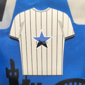 May include: A white and black striped t-shirt design with a blue star featuring a black silhouette of a city skyline. The shirt has short sleeves with black trim and a round neckline. The background is blue.
