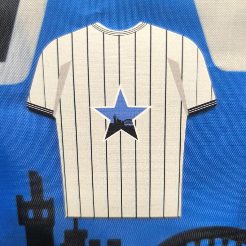 May include: A white and black striped t-shirt design with a blue star featuring a black silhouette of a city skyline. The shirt has short sleeves with black trim and a round neckline. The background is blue.