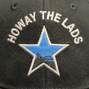 May include: Black baseball cap with white embroidery that reads "Howay The Lads" and a blue star with a city skyline inside.