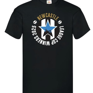 May include: Black t-shirt with a circular design. The design includes a blue star, a cityscape silhouette, and soccer balls. The text reads "NEWCASTLE LEAGUE CUP WINNERS 2025" in gold and white.