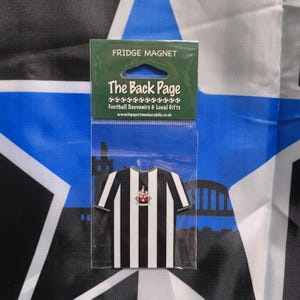 May include: A black and white striped fridge magnet shaped like a soccer jersey with a team crest. The magnet is packaged in a clear plastic bag with the text "The Back Page Football Souvenirs & Local Gifts" and a website address.