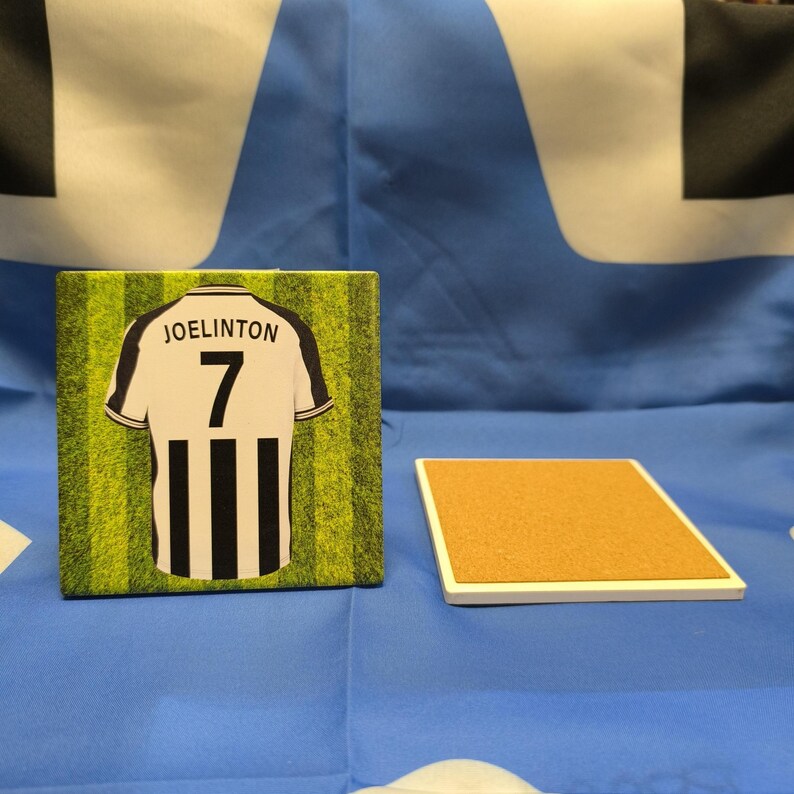 K&ouml;nnte beinhalten: Ein wei&szlig;-schwarzes Fu&szlig;balltrikot mit dem Namen "JOELINGTON" und der Nummer "7" auf der R&uuml;ckseite. Das Trikot befindet sich auf einem gr&uuml;n-wei&szlig;en Hintergrund. Der Untersetzer ist wei&szlig; mit einer Korkoberfl&auml;che.