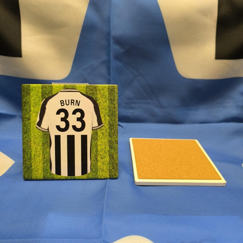 Peut inclure: Sous-verre carr&eacute; avec un motif de maillot de football, affichant "BURN" et le num&eacute;ro "33" sur un fond vert herbeux. Un autre sous-verre carr&eacute; avec une surface en li&egrave;ge est &eacute;galement pr&eacute;sent.