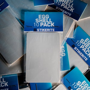 Puede incluir: Una colección de pegatinas rectangulares blancas en envases de plástico transparente. Cada paquete tiene una etiqueta azul con el texto "EGG SHELL 10 PACK" y la marca "STIKERITE".
