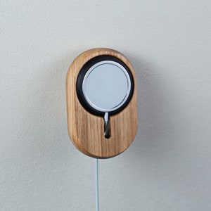 Op de afbeelding: Een houten wandbevestiging voor het opladen van een smartwatch. De houder is gemaakt van lichtbruin hout en heeft een zwarte ronde uitsparing voor de horloge. Een witte oplaadkabel is bevestigd aan de houder.