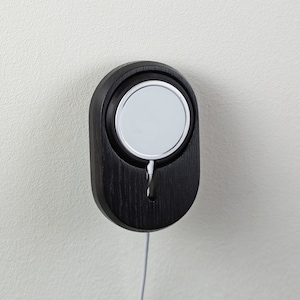 May include: A black wooden wall mount with a white circular charging pad for a smart watch. A white cord hangs from the bottom of the mount.