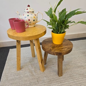 May include: Two wooden stools with a round top and three legs. The stools are made of natural wood with a dark brown stain. A potted plant with green leaves and white flowers sits on the smaller stool. A teapot with a colourful polka dot pattern sits on the larger stool.