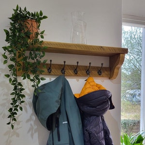 May include: A wooden wall-mounted coat rack with a top shelf and five dark metal hooks. A trailing green plant in a terracotta pot sits on the left side of the shelf, and a clear glass vase stands in the center. Two jackets hang from the hooks.