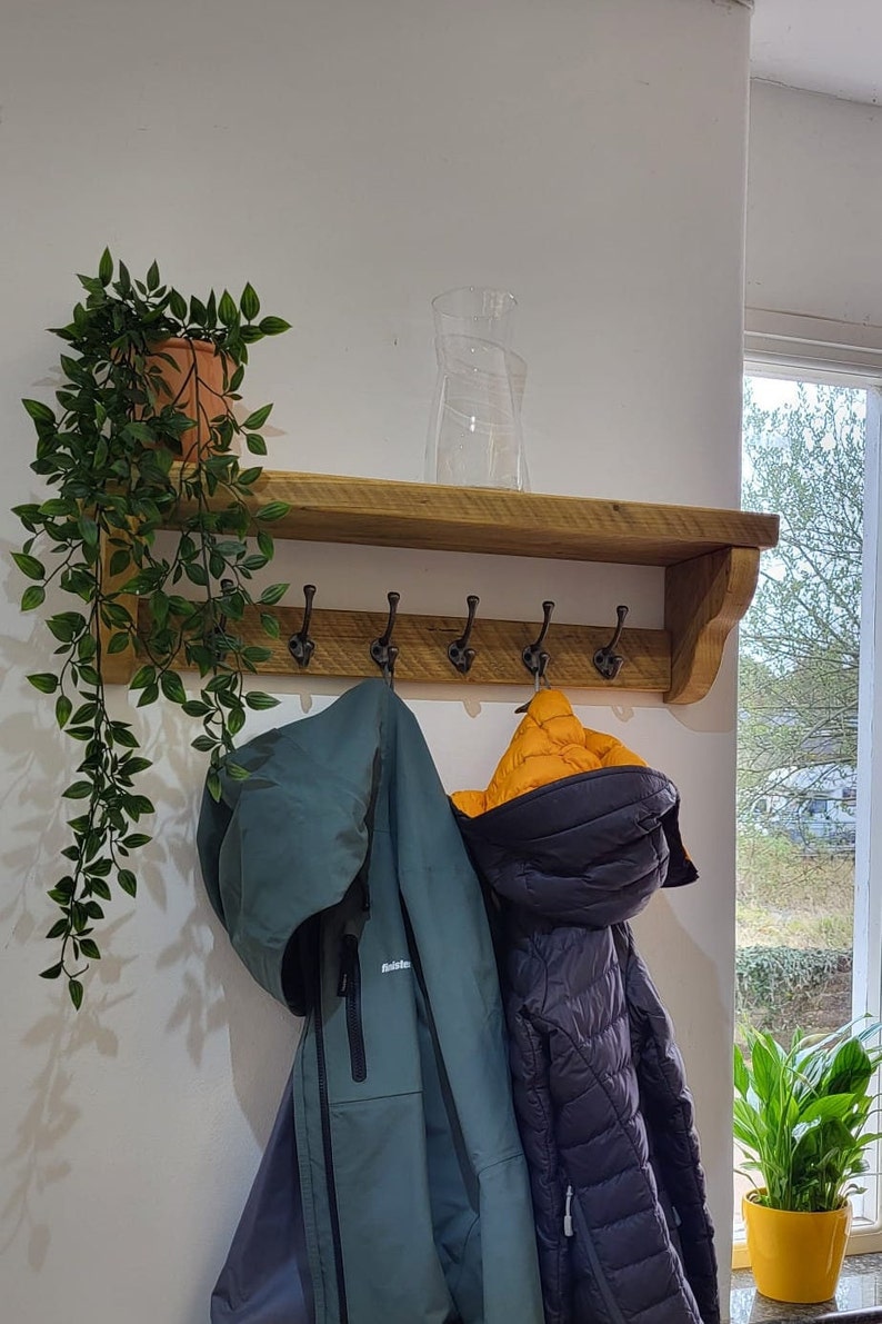May include: A wooden wall-mounted coat rack with a top shelf and five dark metal hooks. A trailing green plant in a terracotta pot sits on the left side of the shelf, and a clear glass vase stands in the center. Two jackets hang from the hooks.