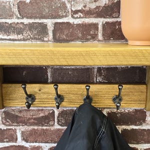 May include: A wall-mounted wooden shelf featuring a smooth, golden-brown finish with two curved side supports. Below the shelf, a horizontal wooden board holds four dark metal hooks, each with a double-pronged design, suitable for hanging items.