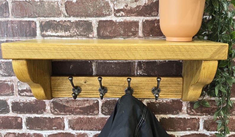 May include: A wall-mounted wooden shelf featuring a smooth, golden-brown finish with two curved side supports. Below the shelf, a horizontal wooden board holds four dark metal hooks, each with a double-pronged design, suitable for hanging items.