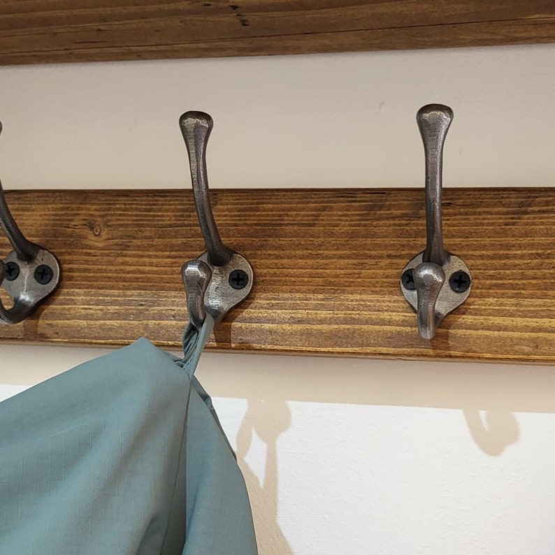 May include: A wall-mounted coat rack featuring a horizontal dark brown stained wooden board with three attached metal hooks. Each hook has a dual-prong design with a brushed silver finish, secured to the wood by two black screws.