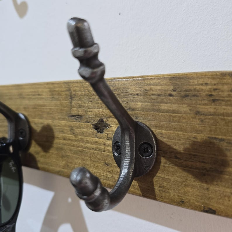 May include: A dark gray metal double hook mounted on a horizontal wooden board. The hook features a curved lower section and a longer, angled upper arm, both with rounded tips. It is secured to the wood with two black screws in a circular base.