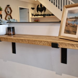 May include: A reclaimed wooden shelf with a natural finish, supported by two black metal brackets. The shelf displays a white potted succulent, a candle in a glass jar, and a framed picture with a gold border. The shelf is mounted on a light grey wall.