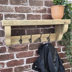 May include: A wall-mounted wooden shelf featuring a flat top surface and a lower horizontal board equipped with four dark metal hooks. The unit is supported by two curved wooden brackets on the sides, with a black jacket hanging from one hook.