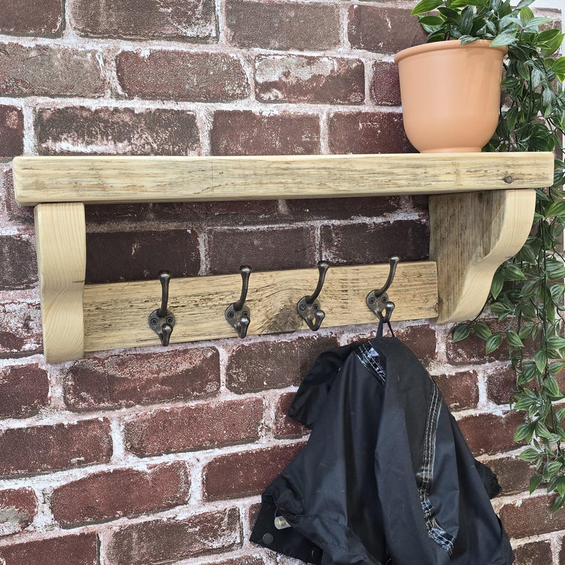 May include: A wall-mounted wooden shelf featuring a flat top surface and a lower horizontal board equipped with four dark metal hooks. The unit is supported by two curved wooden brackets on the sides, with a black jacket hanging from one hook.