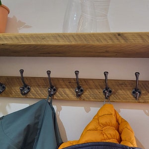May include: A wall-mounted wooden shelf featuring a horizontal plank with six dark metal coat hooks attached. The shelf is constructed from natural-toned wood with visible grain patterns and is supported by two side brackets. The hooks are evenly spaced.