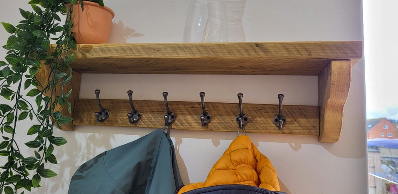 May include: A wall-mounted wooden shelf featuring a horizontal plank with six dark metal coat hooks attached. The shelf is constructed from natural-toned wood with visible grain patterns and is supported by two side brackets. The hooks are evenly spaced.