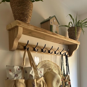 May include: A light brown wooden wall shelf featuring six black metal hooks. The shelf is mounted on a wall and holds two potted plants and a small wooden house ornament. Hanging from the hooks are a beige tote bag, a straw hat, and a patterned bag.