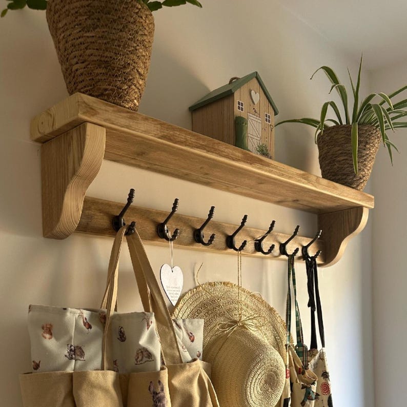 May include: A light brown wooden wall shelf featuring six black metal hooks. The shelf is mounted on a wall and holds two potted plants and a small wooden house ornament. Hanging from the hooks are a beige tote bag, a straw hat, and a patterned bag.
