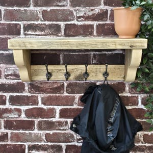 May include: A light-colored wooden wall shelf featuring a flat top surface and a lower horizontal bar equipped with four dark metal hooks. The shelf is supported by two curved wooden brackets on the sides, providing a sturdy structure for storage.