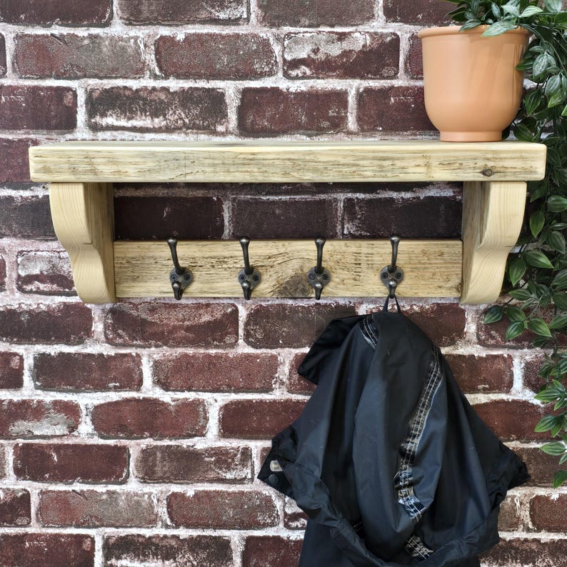 May include: A light-colored wooden wall shelf featuring a flat top surface and a lower horizontal bar equipped with four dark metal hooks. The shelf is supported by two curved wooden brackets on the sides, providing a sturdy structure for storage.