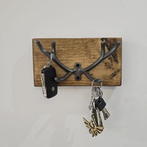 May include: A wooden key holder with a metal antler design. The key holder has three hooks and is mounted on a white wall. Keys and a car key are hanging from the hooks.