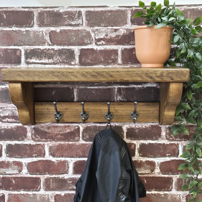 May include: A wall-mounted wooden shelf with a dark oak finish and two side brackets. The unit features a horizontal wooden panel underneath equipped with four dark metal hooks. A black garment hangs from one hook, and a peach-colored pot sits above.