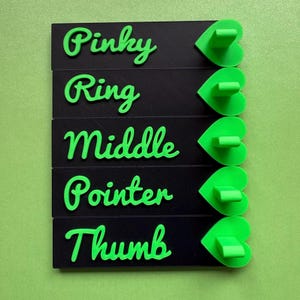 May include: A set of five black finger-shaped ring holders with green heart-shaped bases. Each holder is labeled with a finger name in green cursive: Pinky, Ring, Middle, Pointer, and Thumb. The background is a solid green.