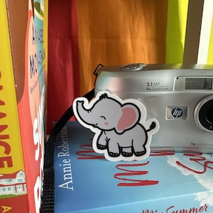 May include: A close-up of a stack of books with a gray elephant sticker on the top book. The book on top is blue with white text that reads "My Mamma Mia Summer". The book below is blue with white text that reads "Annie Robertson". A silver digital camera with the text "2.1 MP 4x digital zoom" is visible in the background.