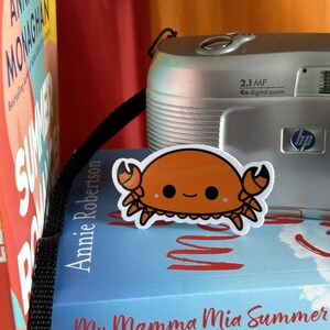 May include: A close-up of a small orange crab sticker with a happy face, placed on top of a blue book with the title "My Mamma Mia Summer". The book is on top of a stack of books, and a silver digital camera with a black strap is visible in the background.