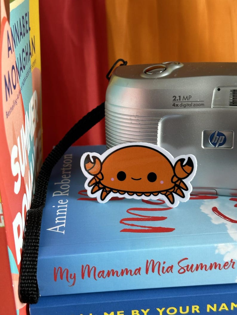 May include: A close-up of a small orange crab sticker with a happy face, placed on top of a blue book with the title "My Mamma Mia Summer". The book is on top of a stack of books, and a silver digital camera with a black strap is visible in the background.