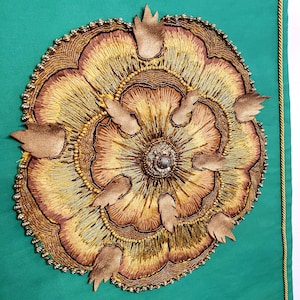 以下が含まれることがあります： ターコイズグリーンの背景に、茶色の芯を持つ大きな金色の刺繍の花。花は銀色のビーズの輪で囲まれています。