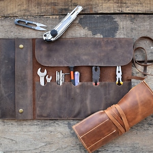 May include: A brown leather tool roll with multiple pockets and a strap closure. The roll is unrolled and contains a variety of tools, including screwdrivers, wrenches, and pliers.
