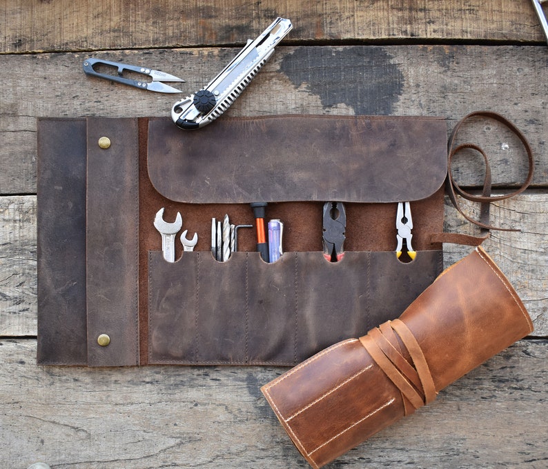 May include: A brown leather tool roll with multiple pockets and a strap closure. The roll is unrolled and contains a variety of tools, including screwdrivers, wrenches, and pliers.